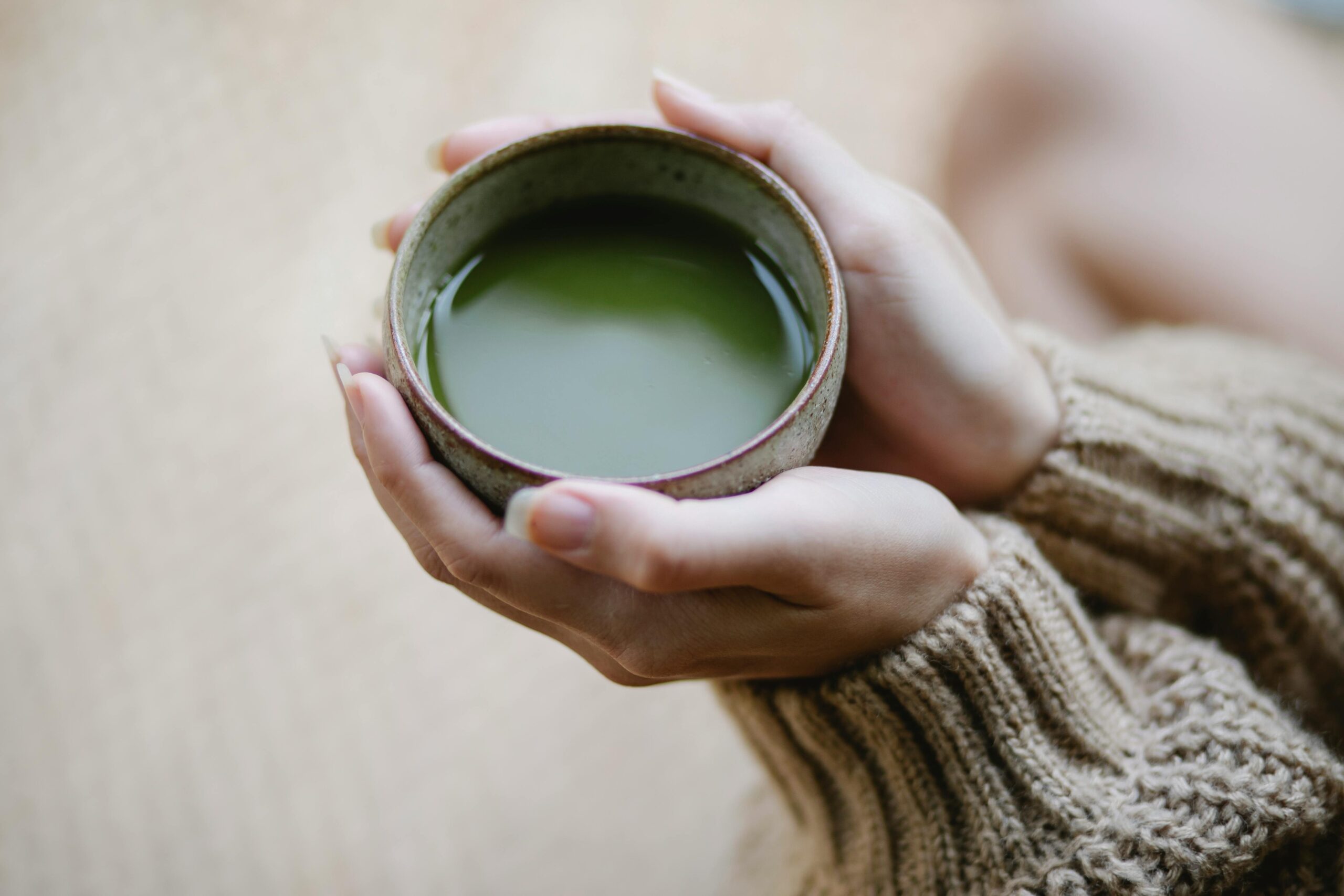 High angle of crop anonymous female in knitwear with ceramic cup of hot matcha tea